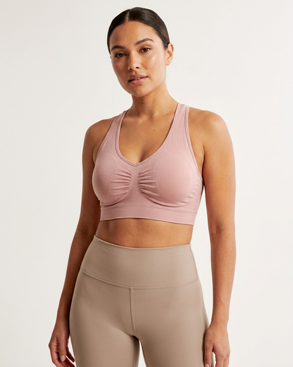 A woman modeling a pink Seamless Racerback Ahh Bra paired with high-waisted beige leggings, against a plain off-white studio background.